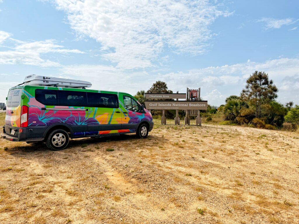 A colorful Escape camper van with vibrant floral artwork is parked on a sandy pull-off next to a wooden sign reading "Gulf Islands National Seashore – Okaloosa Area."