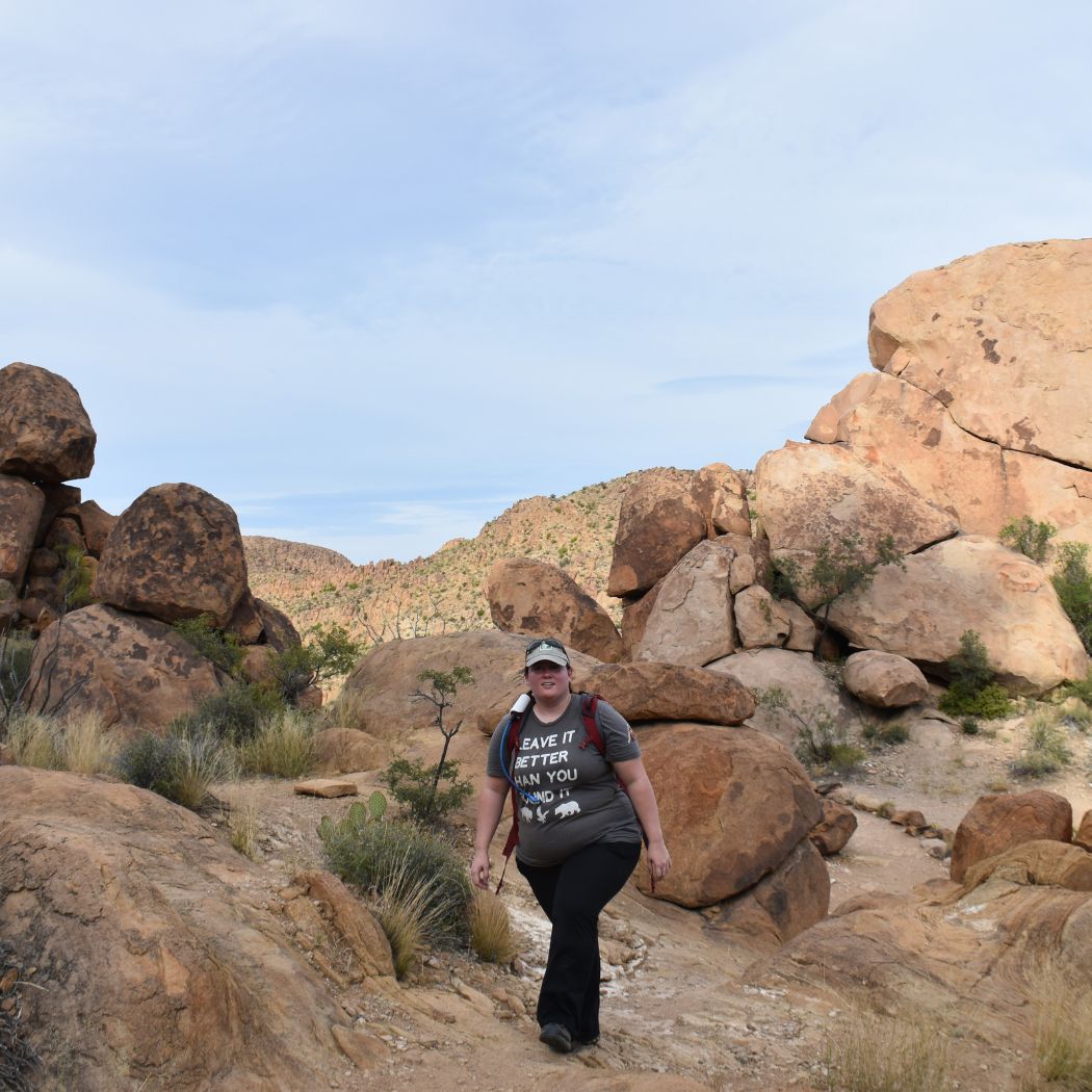 a woman hiking through a rocky desert landscape. She wears a t-shirt that says "LEAVE IT BETTER THAN YOU FOUND IT," emphasizing a message of conservation as she navigates a trail surrounded by large boulders and sparse vegetation under a clear sky.