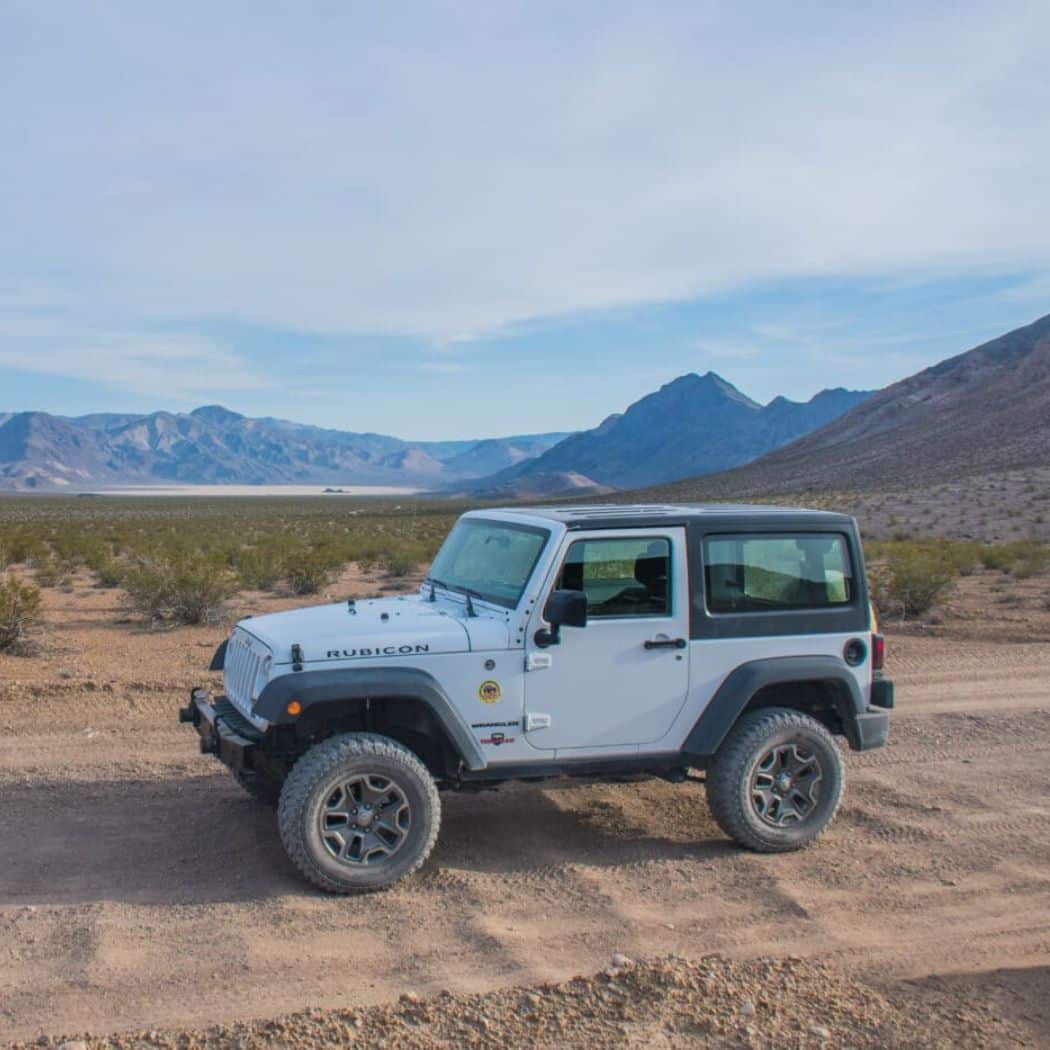 a white Jeep Rubicon on a dirt trail in a desert environment. The jeep is parked with a backdrop of distant mountains under a vast blue sky, ideal for off-road adventures in rugged terrain.