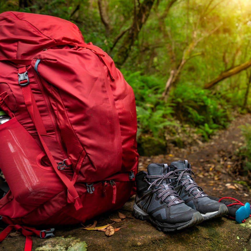 a close-up of a red backpack and a pair of grey hiking boots resting on a rock. The background is a lush green forest bathed in soft sunlight, suggesting a serene hiking or camping scene.