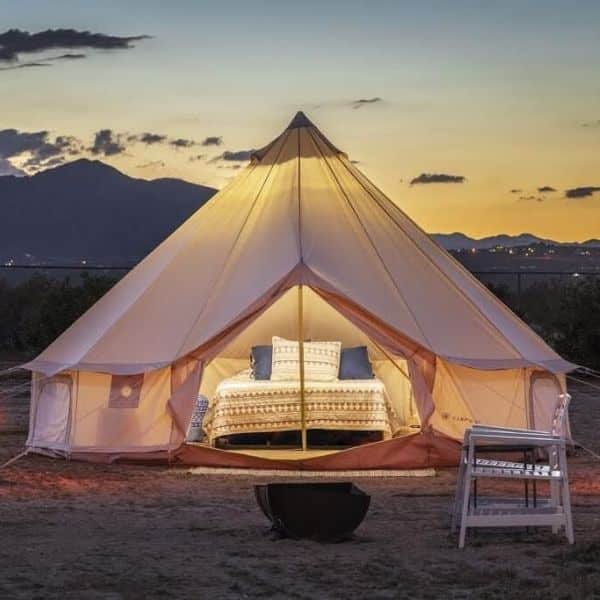 This image features a luxurious, well-lit bell tent set up in a desert landscape at dusk. The tent is open, revealing a comfortable-looking bed with stylish bedding, a large pillow arrangement, and additional seating outside on a small white chair. In the background, soft, early evening light illuminates distant mountains and a hint of city lights, providing a serene, inviting outdoor accommodation experience.