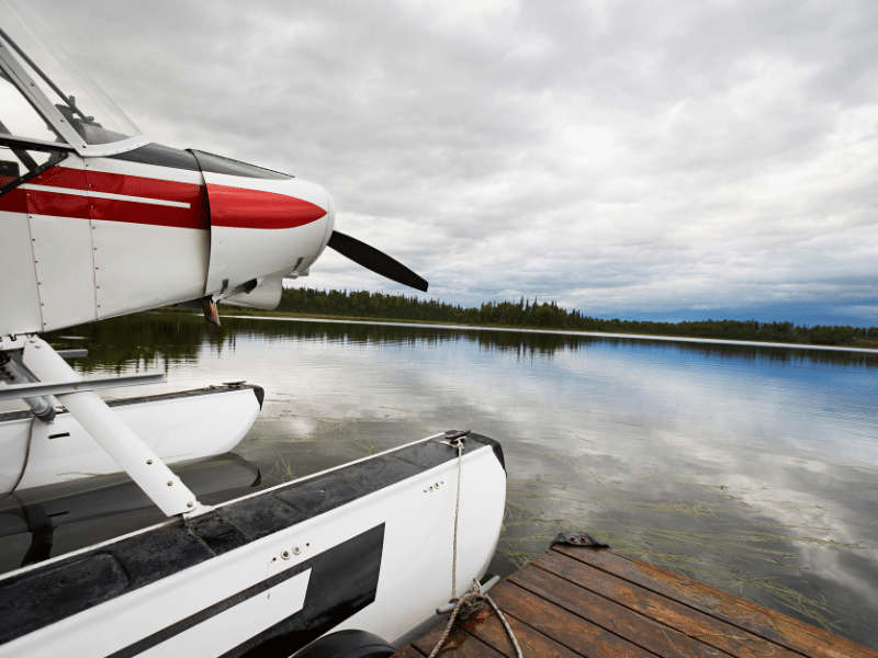 Floatplane docked on a serene Alaskan lake with reflections of surrounding forest and mountains under an overcast sky.