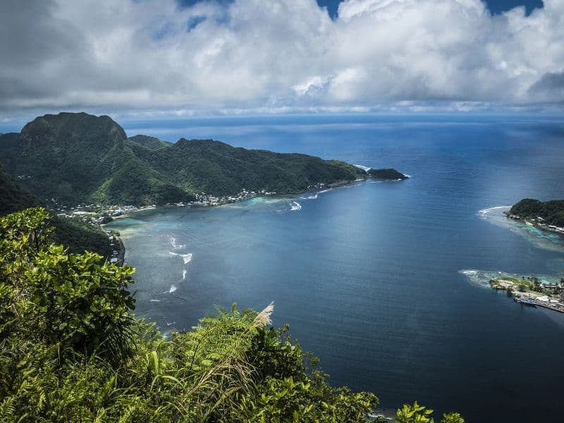 Aerial view of lush green mountains and coastline of the National Park of American Samoa, with a tranquil bay surrounded by dense forest and small settlements.