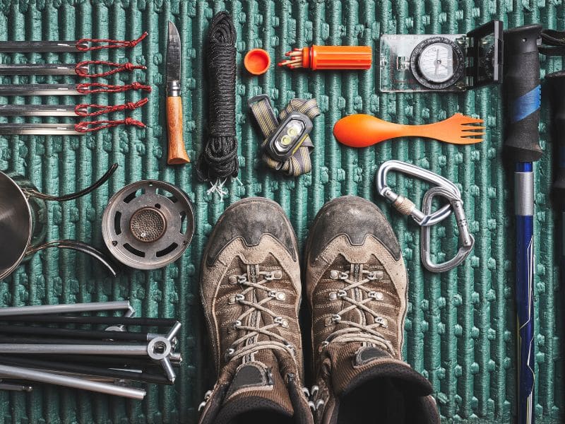 The image features an array of hiking and camping gear neatly arranged on a textured green background. The items include a pair of worn hiking boots, trekking poles, a compass, a camping pot, utensils, and various other tools essential for outdoor adventures. The setup emphasizes preparedness and the joy of engaging with the outdoors.