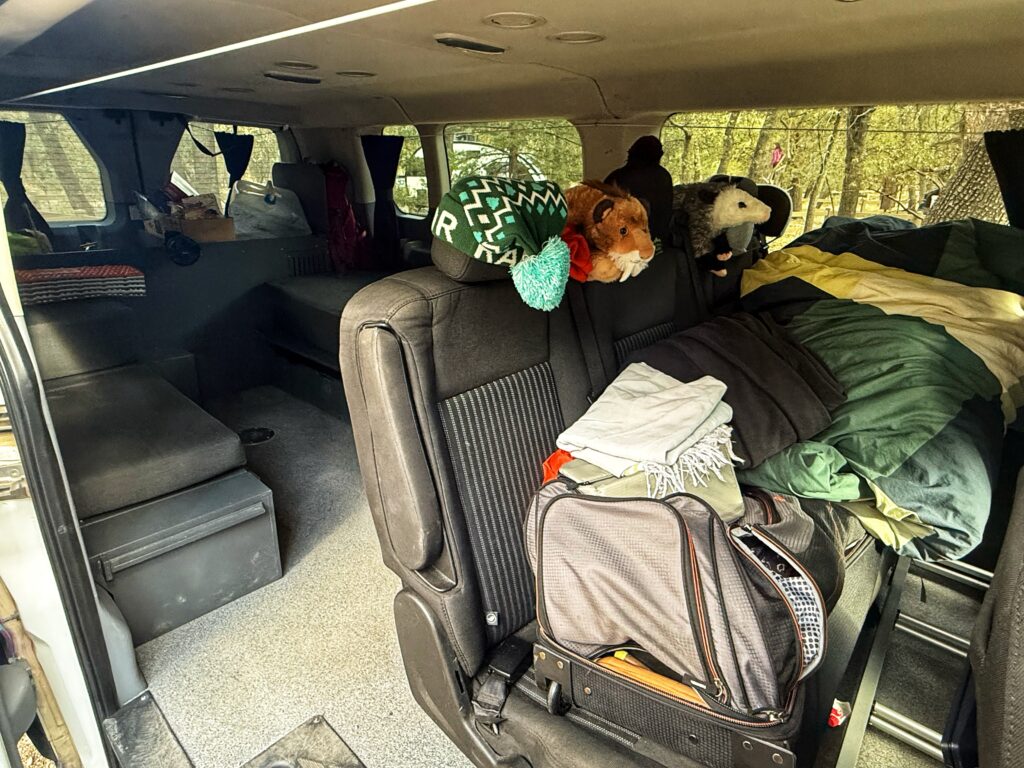The interior of a camper van shows a cozy living space with rear seats partly converted into a bed, covered in green and yellow bedding. A suitcase, folded blankets, and plush animal toys—including a lion and a possum—rest on the seats, while storage bins and supplies are organized in the back, all illuminated by natural light filtering through the windows.