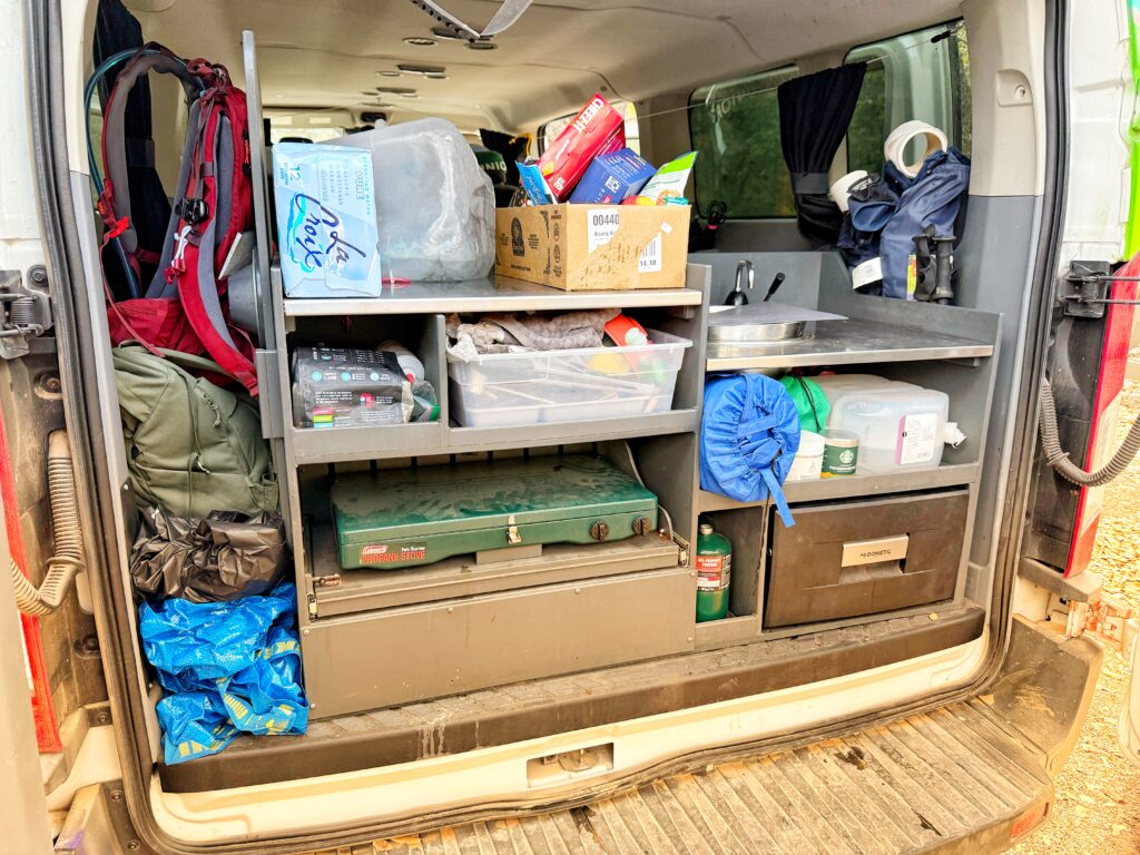The back of a camper van is opened to reveal a neatly organized kitchen and storage area, including a Coleman propane stove, plastic bins of supplies, a sink, water jugs, a red backpack, and a box of La Croix. Various camping essentials like a rolled-up sleeping pad, tape, utensils, and cleaning items are stored on shelves and in cubbies, ready for a road trip or outdoor adventure.