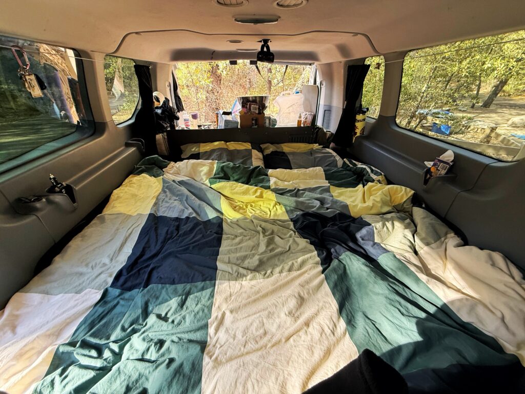 The interior of a camper van is set up with a full-size bed made up in a color-blocked comforter of blues, greens, yellows, and whites. Sunlight filters through the side windows, illuminating the cozy sleeping space, while supplies and snacks are tucked into side compartments and the front storage area.