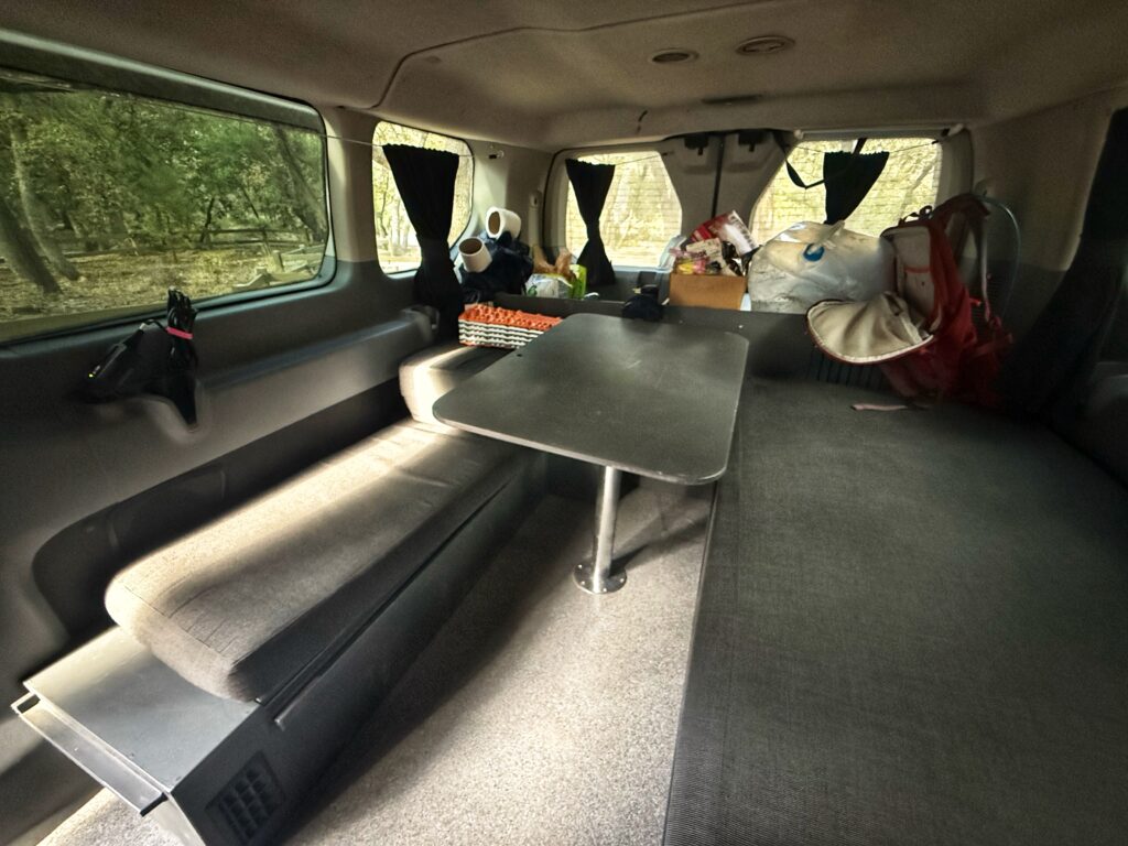 The inside of a camper van is set up in lounge mode with two cushioned benches and a central table, creating a compact dining or work area. Supplies, a red backpack, and a white water jug are neatly stored along the back wall, while windows with dark curtains let in filtered light from the surrounding forest.