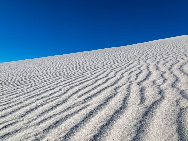 Ripples in white sand dunes under a clear blue sky, creating a striking and minimalistic landscape.
