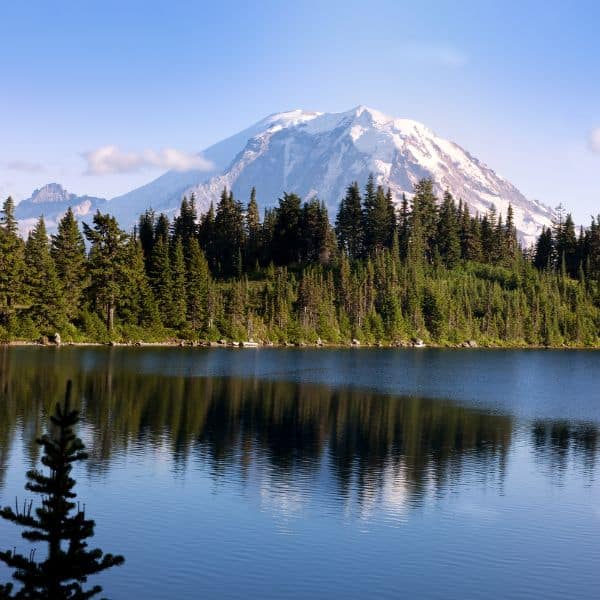 A picturesque mountain with a snow-capped peak, reflected in the calm water of a lake surrounded by evergreen trees. The clear blue sky adds to the serene and pristine beauty of the landscape.