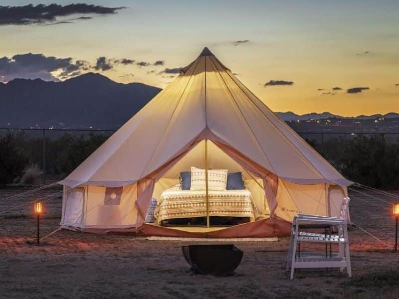 This image features a luxurious, well-lit bell tent set up in a desert landscape at dusk. The tent is open, revealing a comfortable-looking bed with stylish bedding, a large pillow arrangement, and additional seating outside on a small white chair. In the background, soft, early evening light illuminates distant mountains and a hint of city lights, providing a serene, inviting outdoor accommodation experience.