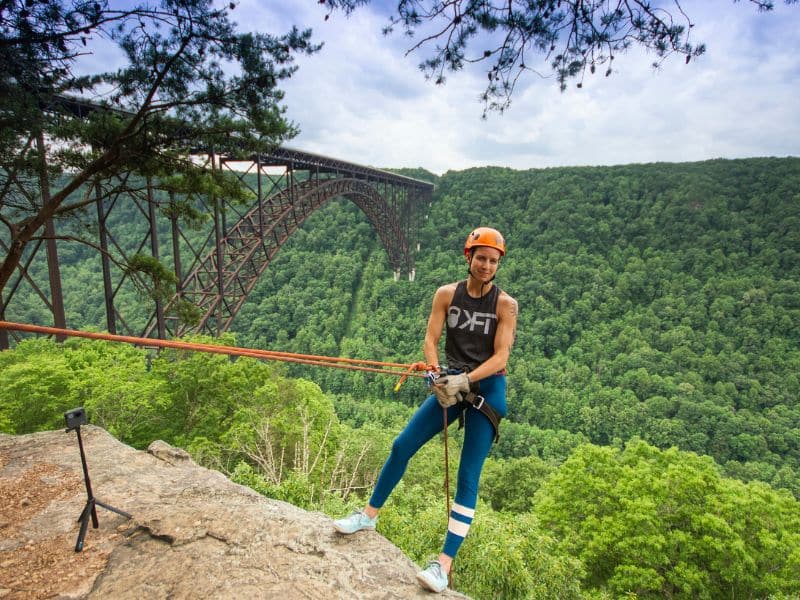 A person standing on a cliff edge with safety ropes, overlooking a forested valley and a bridge in the distance.