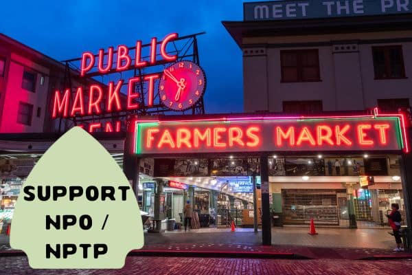 This image features a well-known public market at night, illuminated by bright neon signs reading "PUBLIC MARKET" and "FARMERS MARKET." The background shows the iconic clock, while the foreground displays a green arrow-shaped sign that reads "SUPPORT NPO / NPTP."