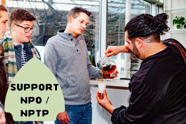 This image depicts a barista pouring coffee for a group of customers in a bright, modern café. The customers look interested and engaged as they watch the barista's demonstration. In the foreground, a green arrow-shaped sign reads "SUPPORT NPO / NPTP."