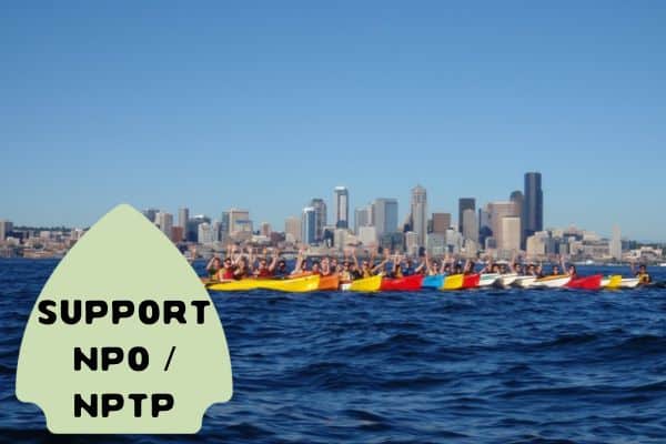 This image shows a group of people kayaking in brightly colored boats on a body of water with a city's skyline in the background under a clear blue sky. In the foreground, a green arrow-shaped sign reads "SUPPORT NPO / NPTP."