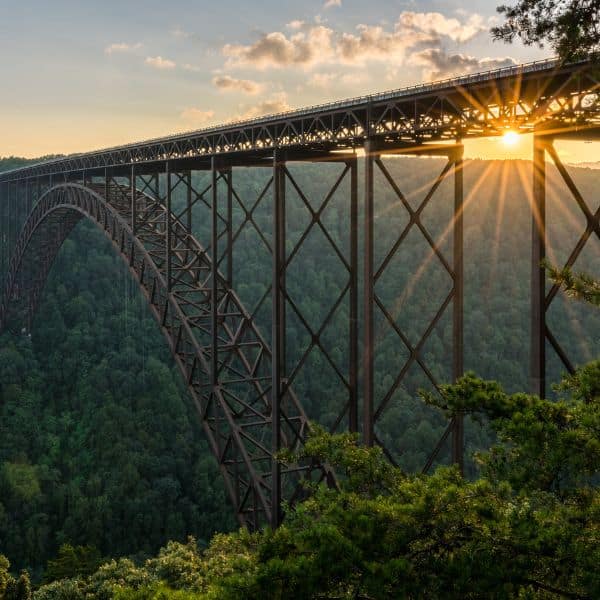 A sunset view of the New River Gorge Bridge with the sun peeking through the steel structure and a forested gorge below.