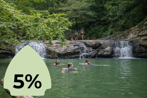 Swimmers enjoying a natural pool at the base of a waterfall surrounded by lush greenery. A green icon with "5%" is superimposed on the left side.