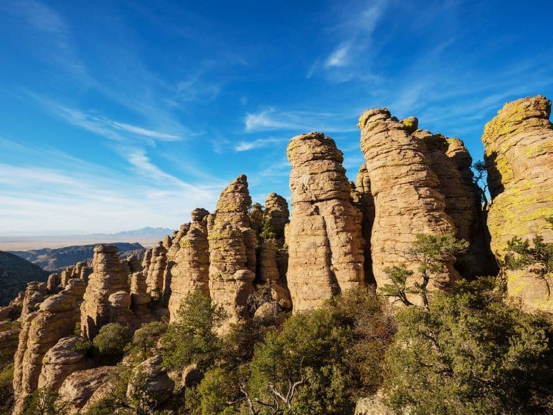 A series of tall, vertical rock formations, also known as hoodoos, set against a bright blue sky with wispy clouds. The hoodoos are characterized by their layered, weathered surfaces and are partially surrounded by green shrubs and trees. The distant horizon reveals a vast, open landscape, highlighting the dramatic scenery.