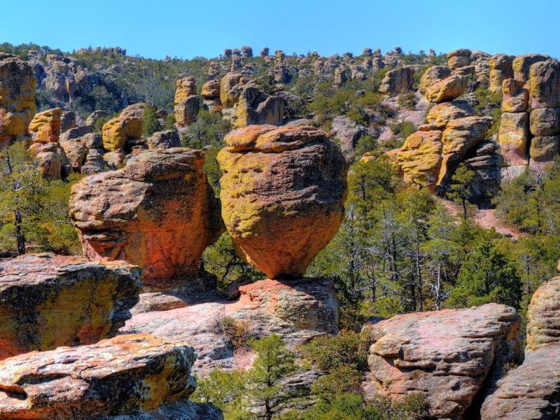 A landscape featuring large, rounded rock formations that balance precariously on narrow bases, surrounded by a backdrop of scattered hoodoos and green vegetation. The distinct orange and yellow hues of the rocks are highlighted by the sunlight, contrasting with the surrounding green trees and the blue sky above.