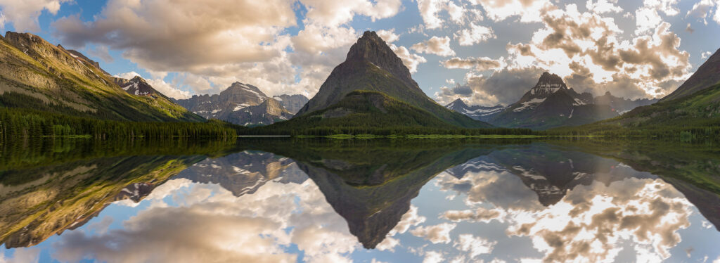 A stunning landscape featuring a range of rugged mountains reflected in a serene lake, with lush green forests and dramatic clouds adding depth to the scene under a partly cloudy sky.