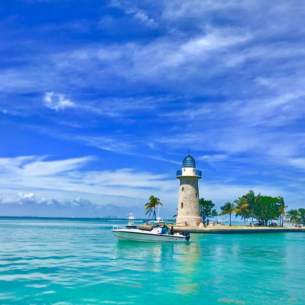 A small boat approaches a historic lighthouse on a palm-lined island in the turquoise waters of Biscayne National Park, under a vibrant blue sky.