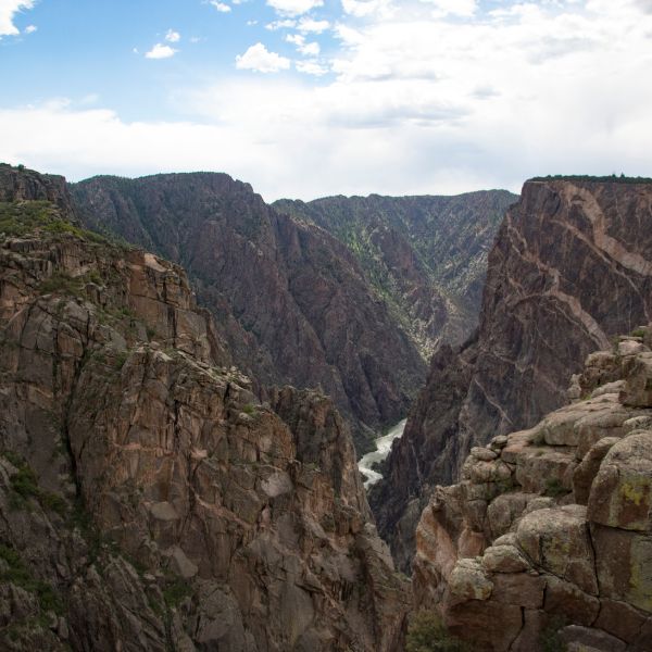 Steep canyon walls tower over the narrow, winding Gunnison River below in Black Canyon of the Gunnison National Park, with clouds scattered across the sky.