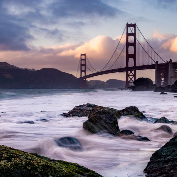 A dramatic view of the Golden Gate Bridge at sunset, with waves crashing against coastal rocks and clouds glowing with soft golden light.