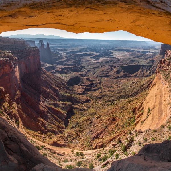 A panoramic view of Canyonlands National Park seen through a rock arch, with a vast desert canyon stretching to the horizon under a hazy sky.