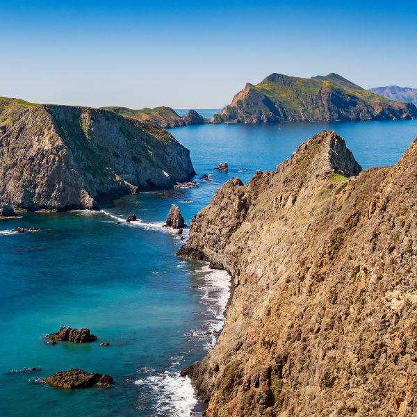 The Channel Islands rise from the Pacific Ocean, with jagged cliffs, rocky shores, and lush green hilltops bathed in sunlight on a calm day.