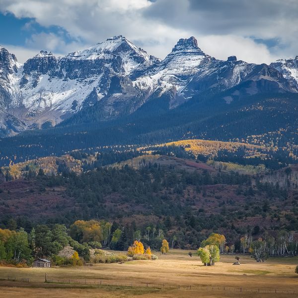 A breathtaking mountain scene in Colorado featuring snow-capped peaks, dense pine forests, and golden autumn trees across a wide, open meadow.