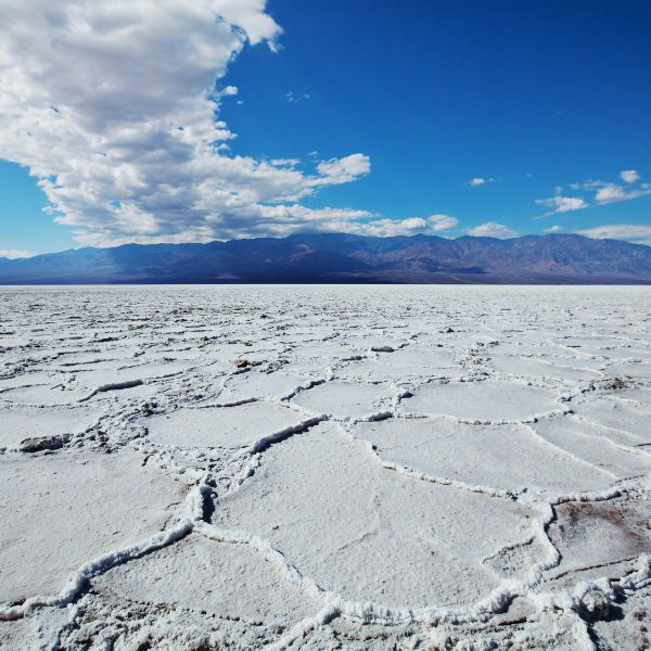 Salt flats in Death Valley National Park create a cracked, geometric pattern across a stark white desert floor, with distant mountains under a dramatic sky.