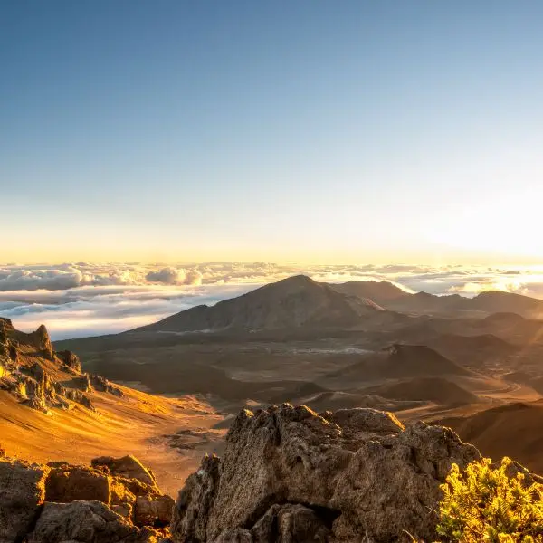 The sun dips below the horizon, casting golden light over the Haleakalā crater. Rocky formations in the foreground add depth to the scene, while a layer of clouds blankets the distant mountains.