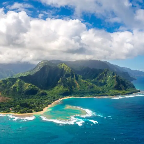 An aerial view of a stunning Hawaiian coastline with lush green mountains meeting the turquoise ocean. White waves crash against the golden sandy shore, with soft clouds hovering over the peaks.