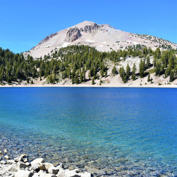 Clear alpine lake at the base of a rocky, tree-covered mountain in Lassen Volcanic National Park, with a vibrant blue sky and evergreen trees reflecting in the water.