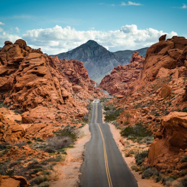 A two-lane road winding through dramatic red rock formations in the Nevada desert, leading toward a distant mountain under a partly cloudy sky.