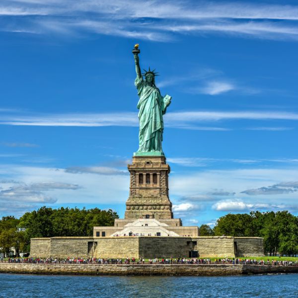 A bright, clear view of the Statue of Liberty standing tall on Liberty Island in New York Harbor, surrounded by greenery and visitors along the base.