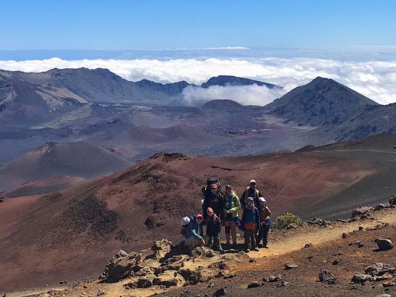 A group of hikers, dressed in outdoor gear and carrying backpacks, poses on a rocky trail in a volcanic landscape. The terrain consists of red and black lava rock, with rugged peaks in the background and a layer of clouds below the mountain ridges. The sky is clear blue, suggesting high-altitude conditions.