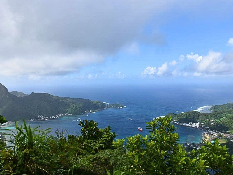 A lush, green coastline curves around a deep blue bay under a partly cloudy sky, with small boats visible in the water and clusters of buildings dotting the shore. Dense tropical foliage fills the foreground, adding depth to the scenic, panoramic view of the coastal landscape.