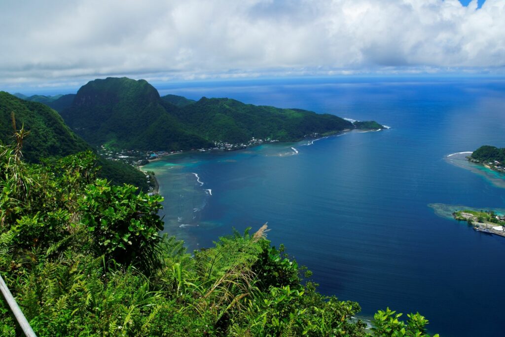 Sweeping aerial view of American Samoa’s coastline, with lush green peaks, turquoise reef waters, and white ocean surf curving around the bays.