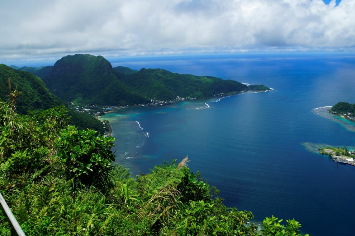Sweeping aerial view of American Samoa’s coastline, with lush green peaks, turquoise reef waters, and white ocean surf curving around the bays.