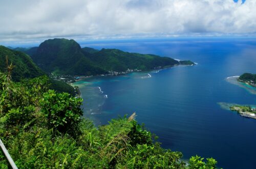 Sweeping aerial view of American Samoa’s coastline, with lush green peaks, turquoise reef waters, and white ocean surf curving around the bays.