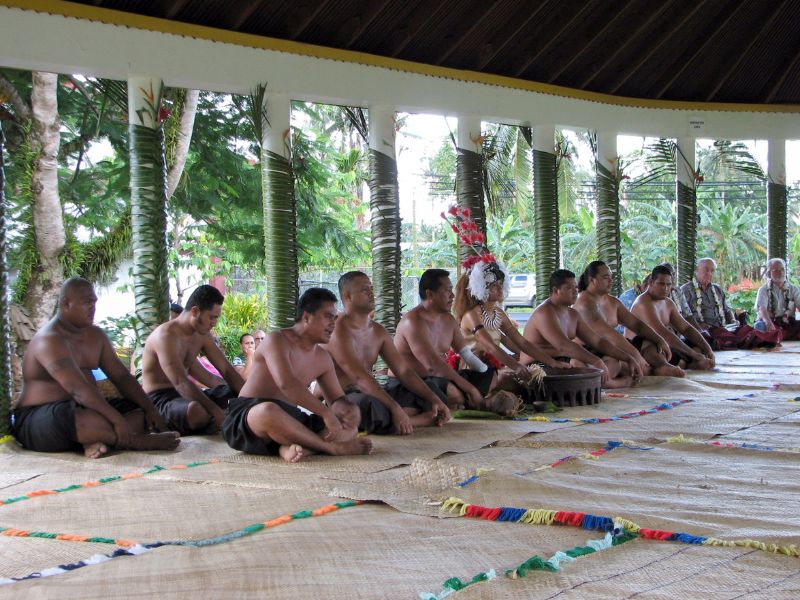 A group of Samoan men dressed in traditional attire sit cross-legged on woven mats inside a fale, a traditional open-sided meeting house. A woman in ceremonial dress with a feathered headdress sits at the center, surrounded by others participating in a cultural gathering or ceremony.