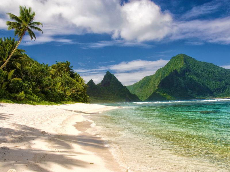A pristine white sand beach lined with palm trees stretches along turquoise waters with dramatic green mountains in the background on Ofu Island in American Samoa. Gentle waves lap the shore under a partly cloudy sky, creating a serene tropical scene.