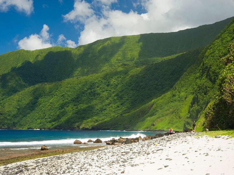 Lush green cliffs rise steeply behind a white sand and rocky beach on the island of Ta‘ū in American Samoa. Two hikers walk along the shoreline near the edge of the forest, with turquoise ocean waves gently crashing against the rocks.