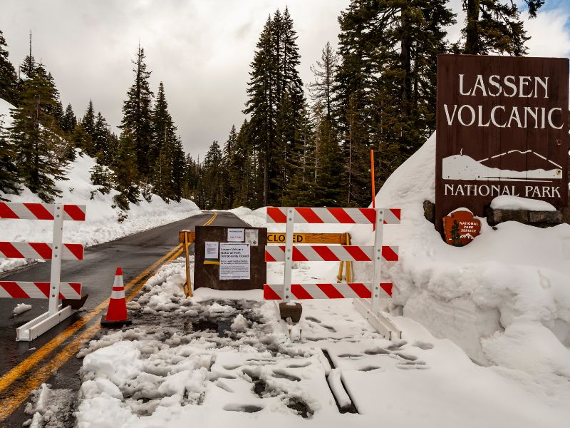 A snowy road into Lassen Volcanic National Park is blocked by striped barricades, cones, and closure notices posted on a wooden board. A large park entrance sign stands to the right, surrounded by deep snow.