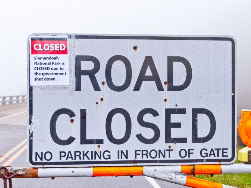 A large road sign reads “ROAD CLOSED – NO PARKING IN FRONT OF GATE,” with a smaller sign above stating Shenandoah National Park is closed due to a government shutdown. The scene is foggy with part of a guardrail and road visible behind the sign.