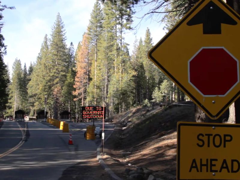 A forest road is blocked off with a digital sign reading “DUE TO GOVERNMENT SHUTDOWN” in orange lights. A yellow traffic sign in the foreground warns "STOP AHEAD" with an upward arrow.