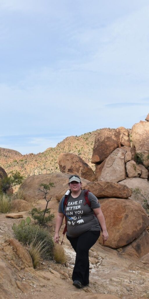 Person hiking on a rocky desert trail surrounded by large boulders and dry brush under a pale blue sky. They are wearing a gray T shirt that reads Leave It Better Than You Found It with animal silhouettes along the bottom along with a baseball cap backpack and black pants. The image highlights responsible outdoor adventure and exploring rugged desert landscapes.