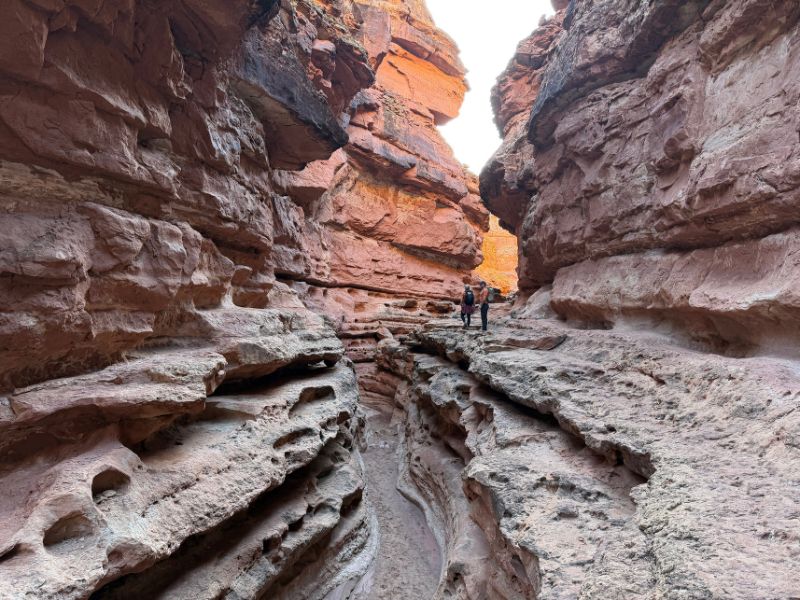 Two hikers explore a dramatic, carved sandstone canyon with layered rock formations and glowing light. The photo captures a memorable moment on a Pronghorn Expeditions Glen Canyon hiking tour.