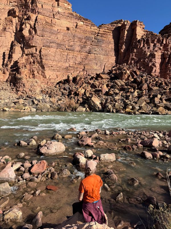 Person in an orange shirt stands at the rocky edge of a flowing river with towering red canyon walls behind. Scene captures the raw beauty and remote nature of a Glen Canyon hiking tour with Pronghorn Expeditions.